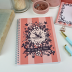 A wire bound A5 notebook with pink striped cover and purple floral illustrations, and the text "All of my Best Ideas", on a white desk with stationery items