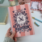 A hand holding a wire bound A5 notebook with pink striped cover and purple floral illustrations, and the text "All of my Best Ideas"
