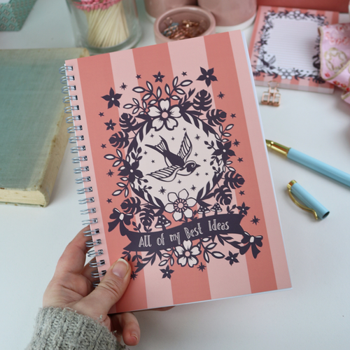 A hand holding a wire bound A5 notebook with pink striped cover and purple floral illustrations, and the text "All of my Best Ideas"