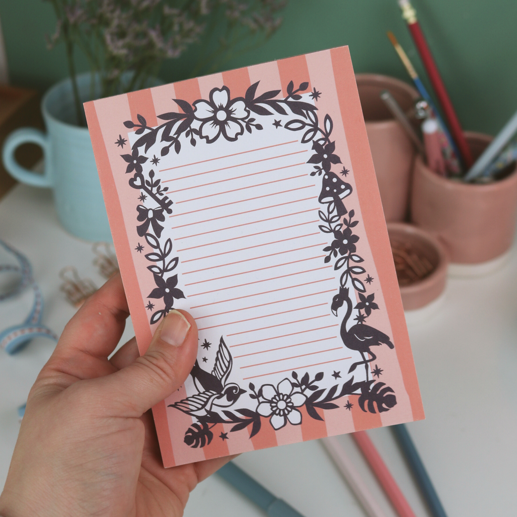 Hand holding a decorative notepad with floral and bird designs on a desk.