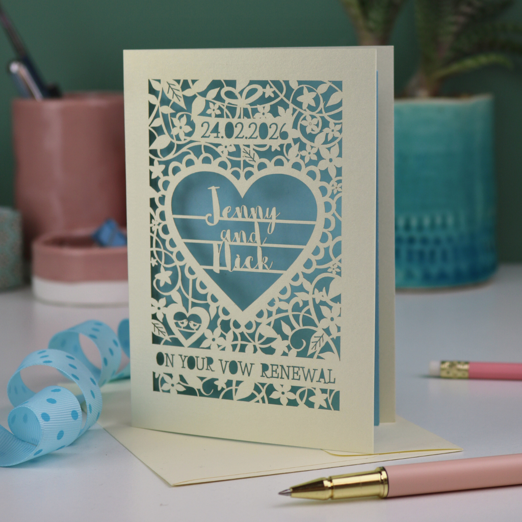Laser-cut vow renewal card with personalized text on a table with stationery items.