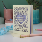 Laser-cut card for a vow renewal with personalized text on a desk with stationery items.