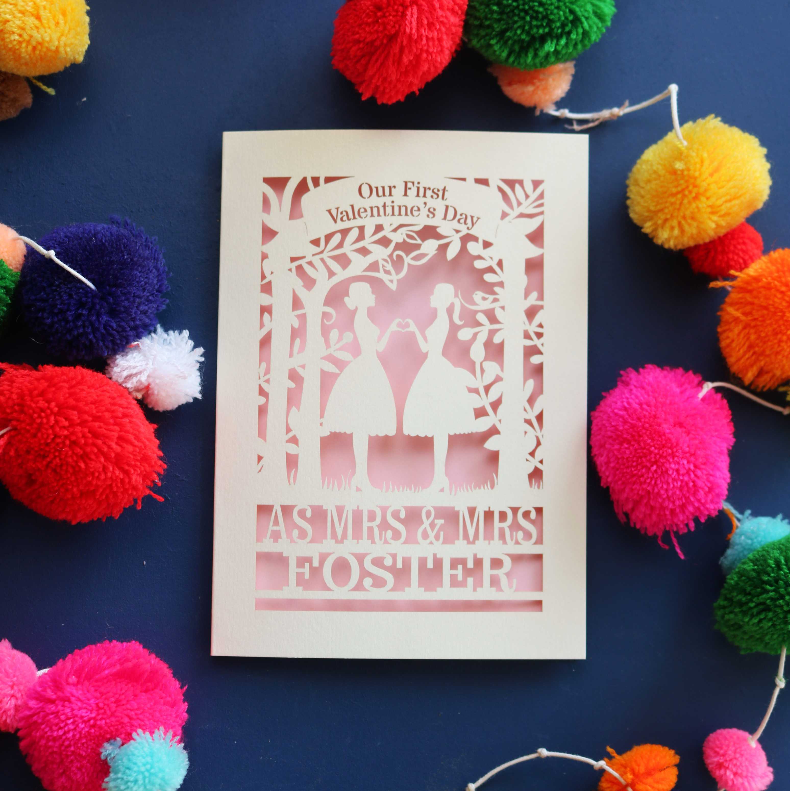 A personalised Valentine's day card with a laser cut design of two women making a heart shape, with a blank space for a personal message, and brightly coloured pom-pom decorations around it.