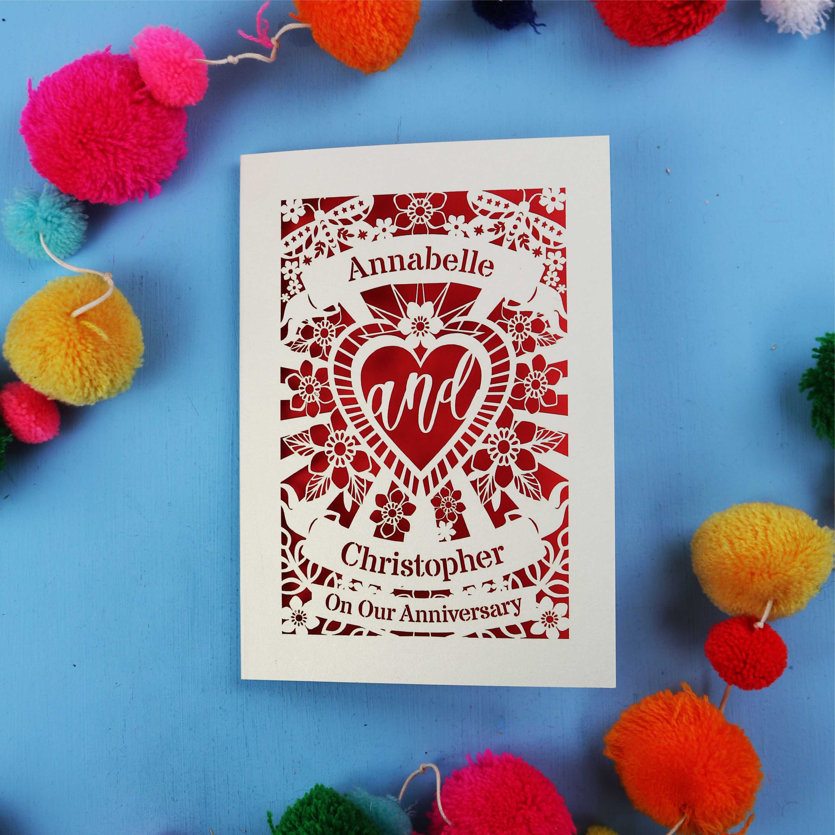 Personalised laser cut anniversary card with decorative heart design on a blue background with colourful pom-poms.