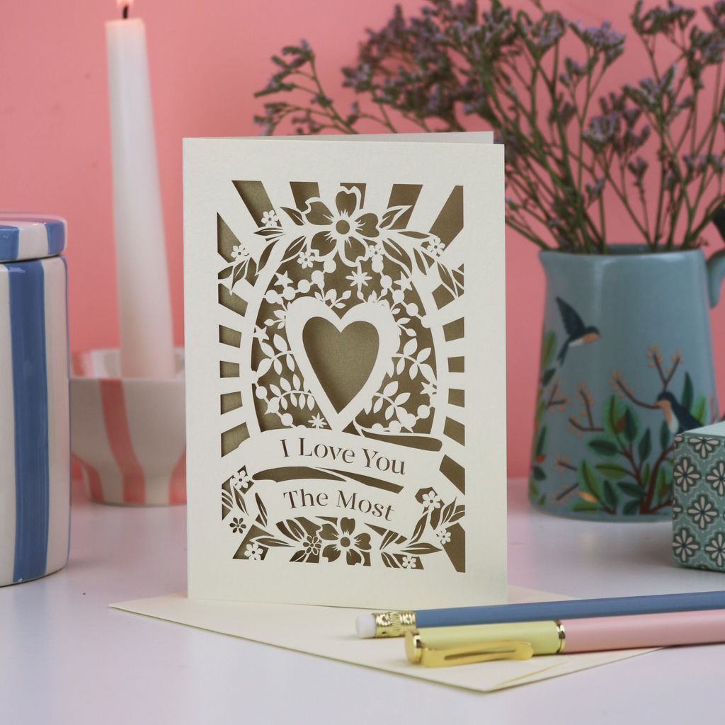 A paper cut Valentine's card that says "I love you the most", with heart design and gold paper insert. Card sits on a desk with stationery items in front of a pink background