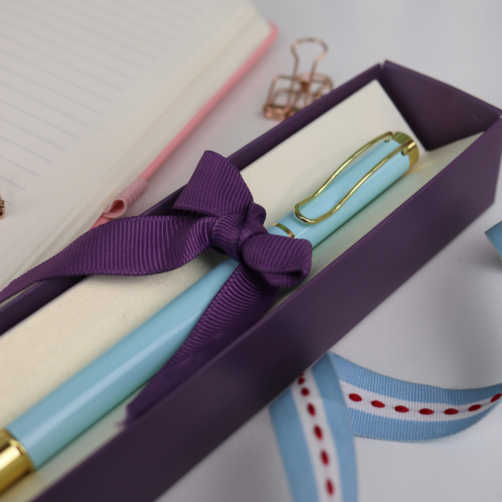 Light blue pen with gold accents in a purple box, tied with a purple ribbon, next to a pink notebook.