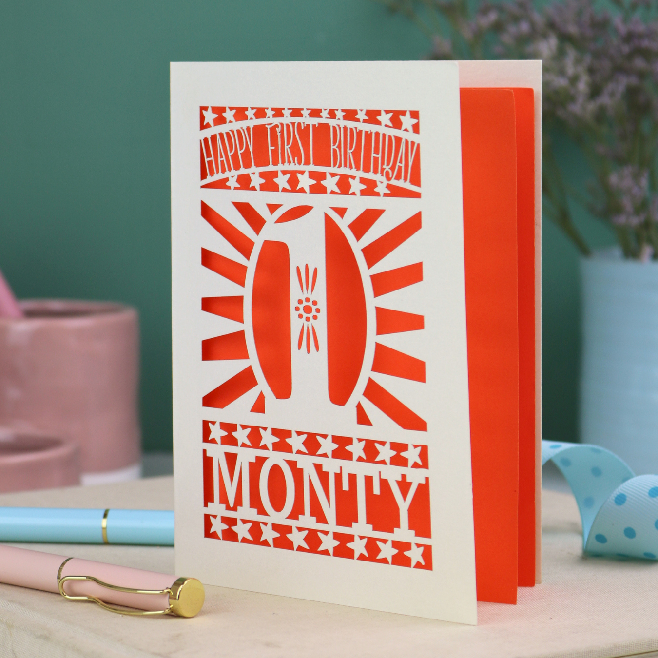 Personalised Baby's 1st Birthday card. laser cut with star borders and "Happy First Birthday" at the top, a decorative cut out number 1 in the centre and personalised with the name "MONTY" at the bottom. card is cream with a bright orange paper backing
