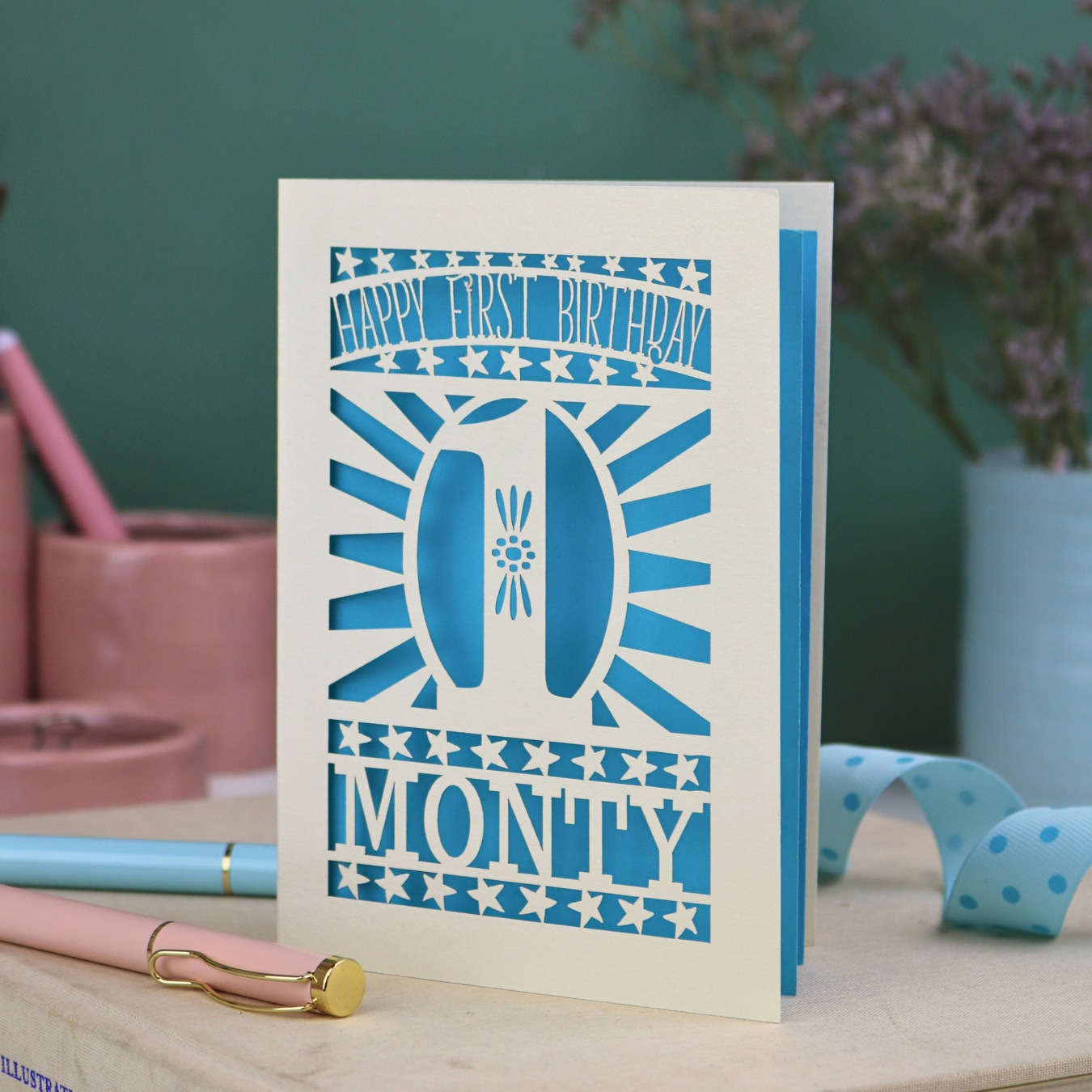Personalised Baby's 1st Birthday card. laser cut with star borders and "Happy First Birthday" at the top, a decorative cut out number 1 in the centre and personalised with the name "MONTY" at the bottom. card is cream with a bright blue paper backing