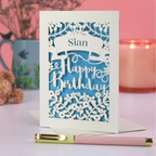 A laser cut floral birthday card with a name and the words "Happy Birthday", on white desk with pink background.