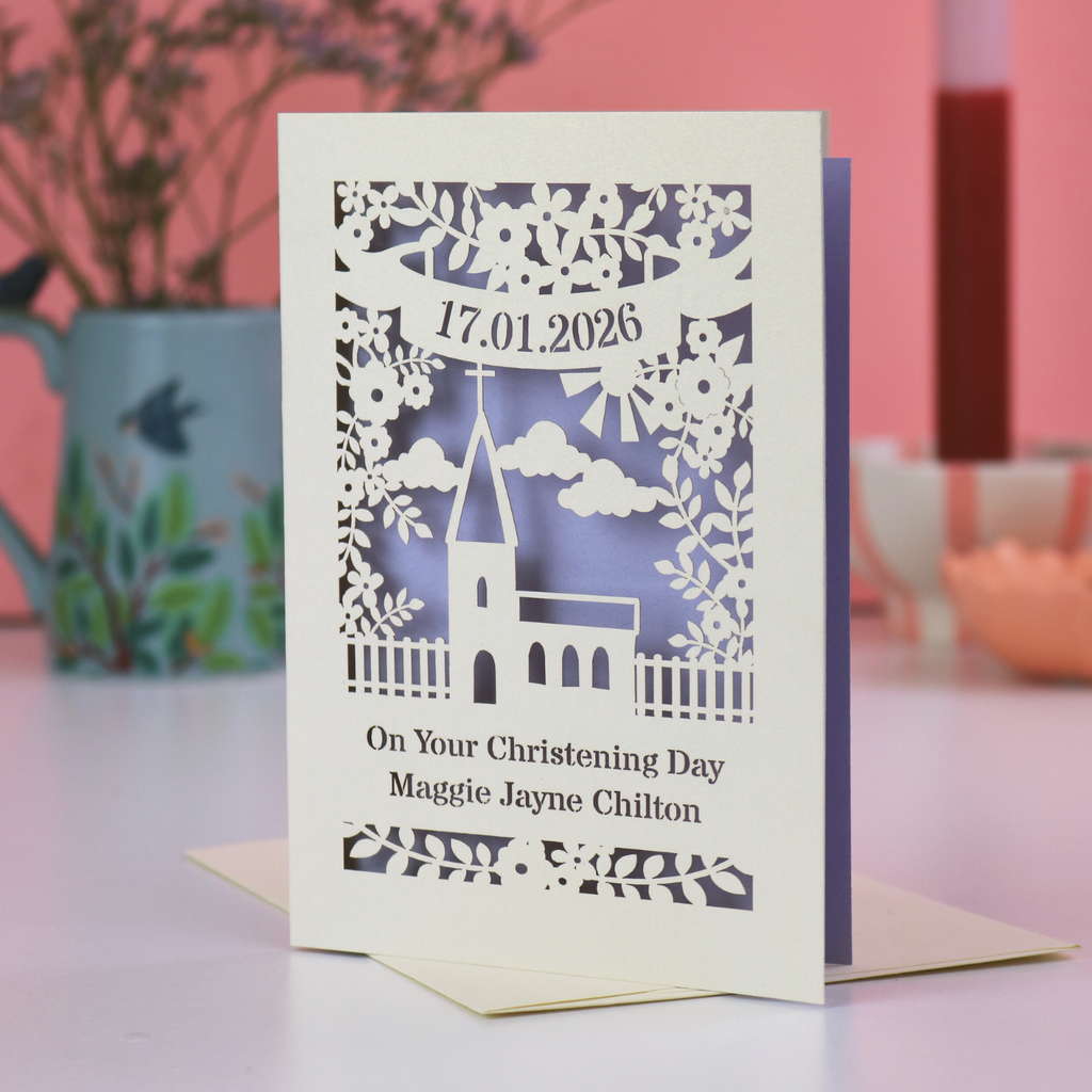 A laser cut Christening card with church design, on a white desk with pink background