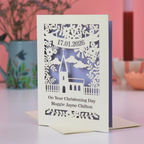 A laser cut Christening card with church design, on a white desk with pink background