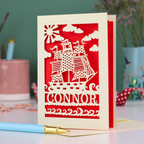 A laser cut boys birthday card with cut out pirate ship design, personalised with the name "Connor". Card stands on a white desk with a pen and stationery items