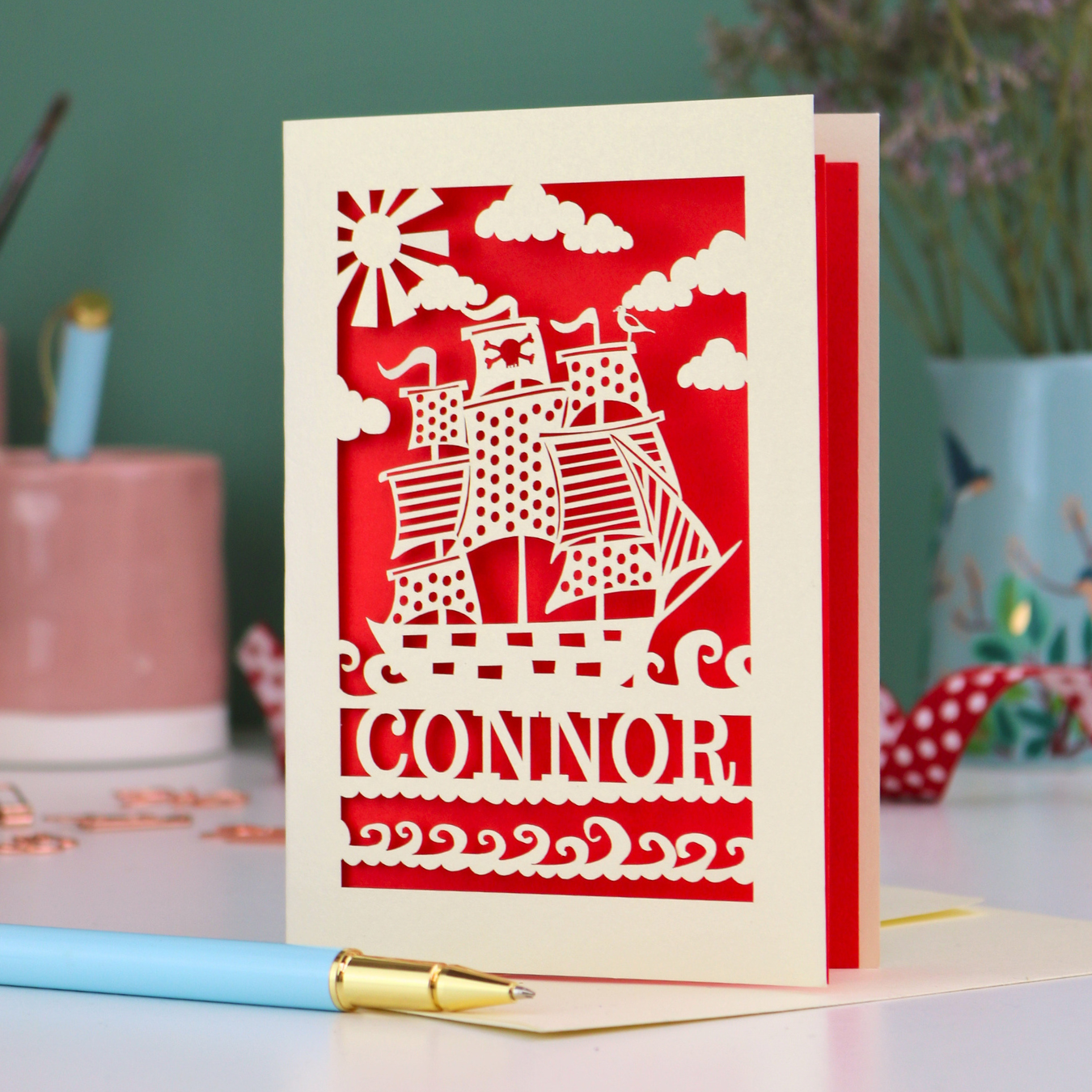 A laser cut boys birthday card with cut out pirate ship design, personalised with the name "Connor". Card stands on a white desk with a pen and stationery items