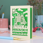 A laser cut boys birthday card with cut out pirate ship design, personalised with the name "Connor". Card stands on a white desk with a pen and stationery items
