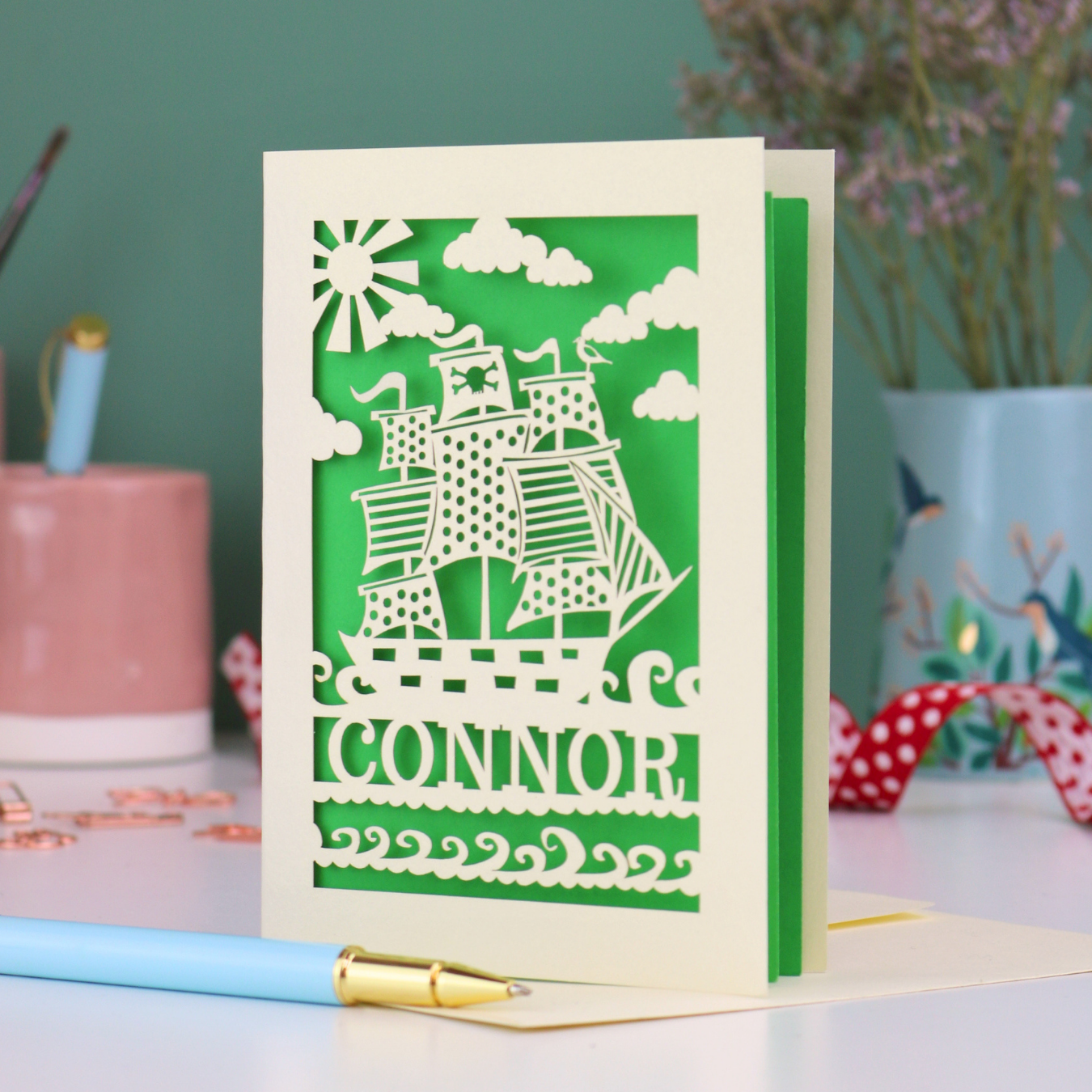 A laser cut boys birthday card with cut out pirate ship design, personalised with the name "Connor". Card stands on a white desk with a pen and stationery items