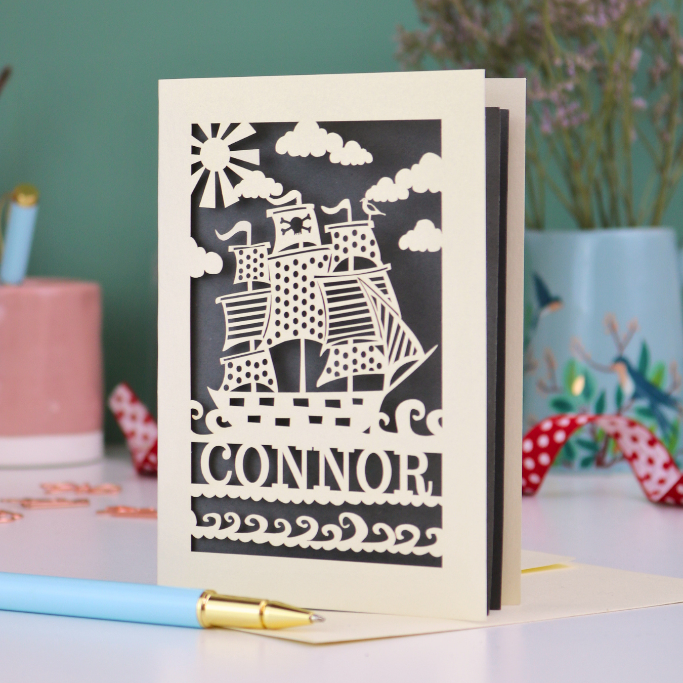 A laser cut boys birthday card with cut out pirate ship design, personalised with the name "Connor". Card stands on a white desk with a pen and stationery items