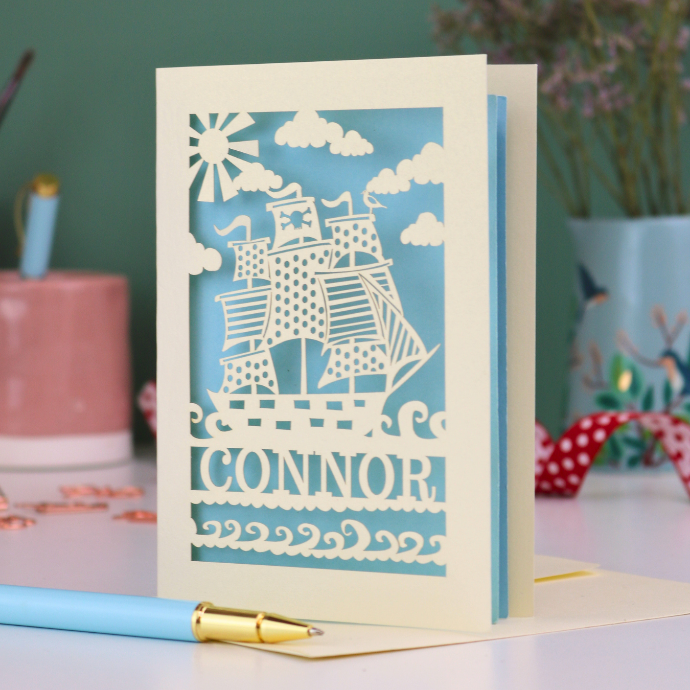 A laser cut boys birthday card with cut out pirate ship design, personalised with the name "Connor". Card stands on a white desk with a pen and stationery items