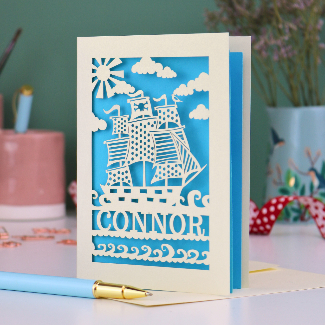 A laser cut boys birthday card with cut out pirate ship design, personalised with the name "Connor". Card stands on a white desk with a pen and stationery items