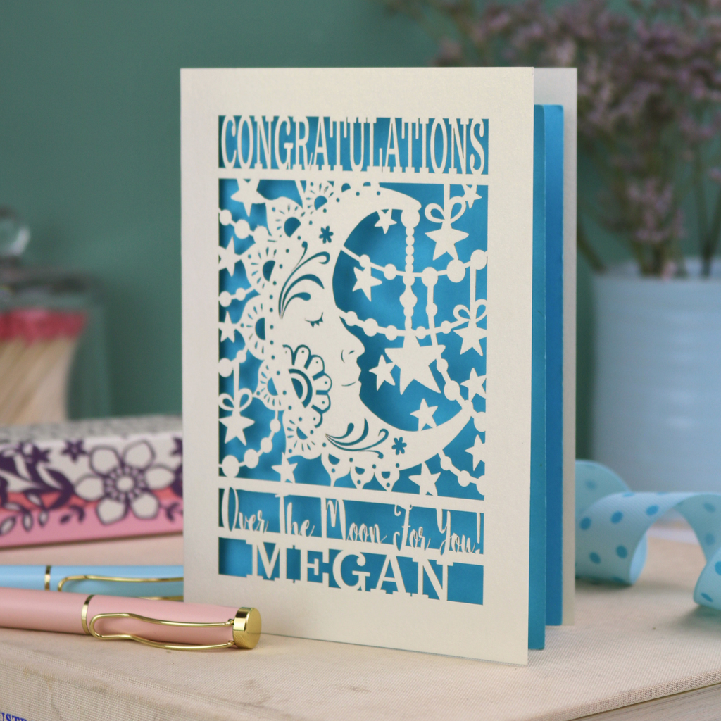 Personalised Congratulations Card with laser cut crescent moon design and text 'Congratulations, Over the moon for you Megan' on a wooden surface.