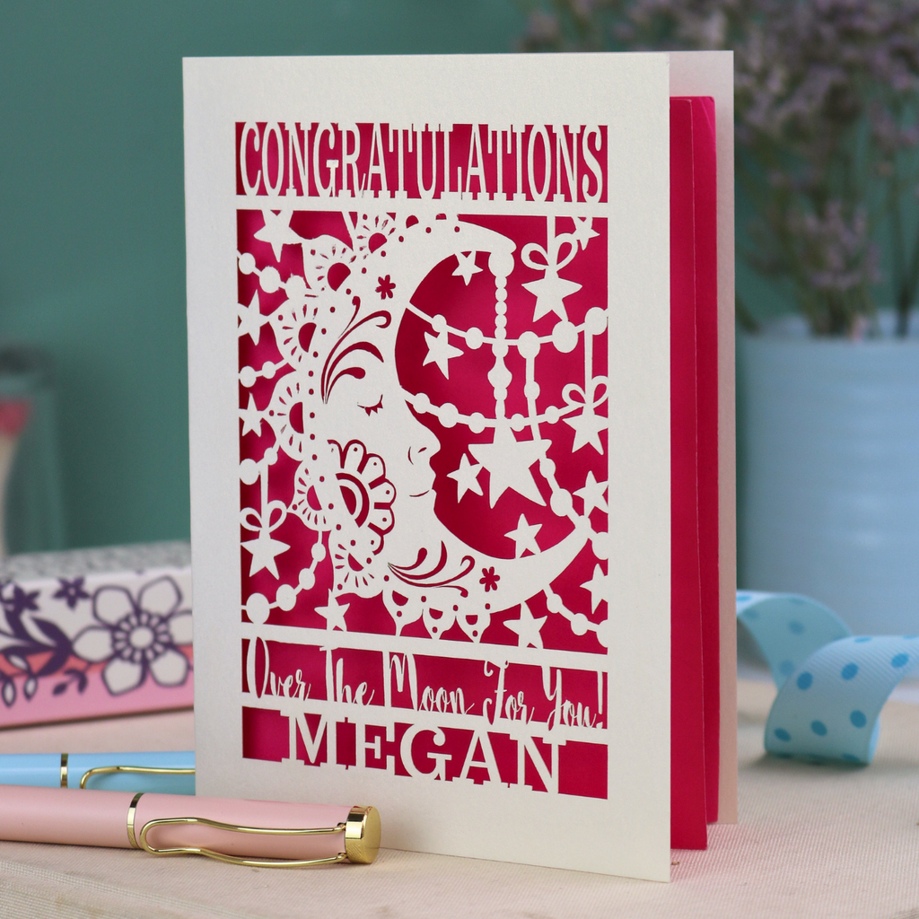 Personalised Congratulations Card with laser cut crescent moon design and text 'Congratulations, Over the moon for you Megan' on a wooden surface.