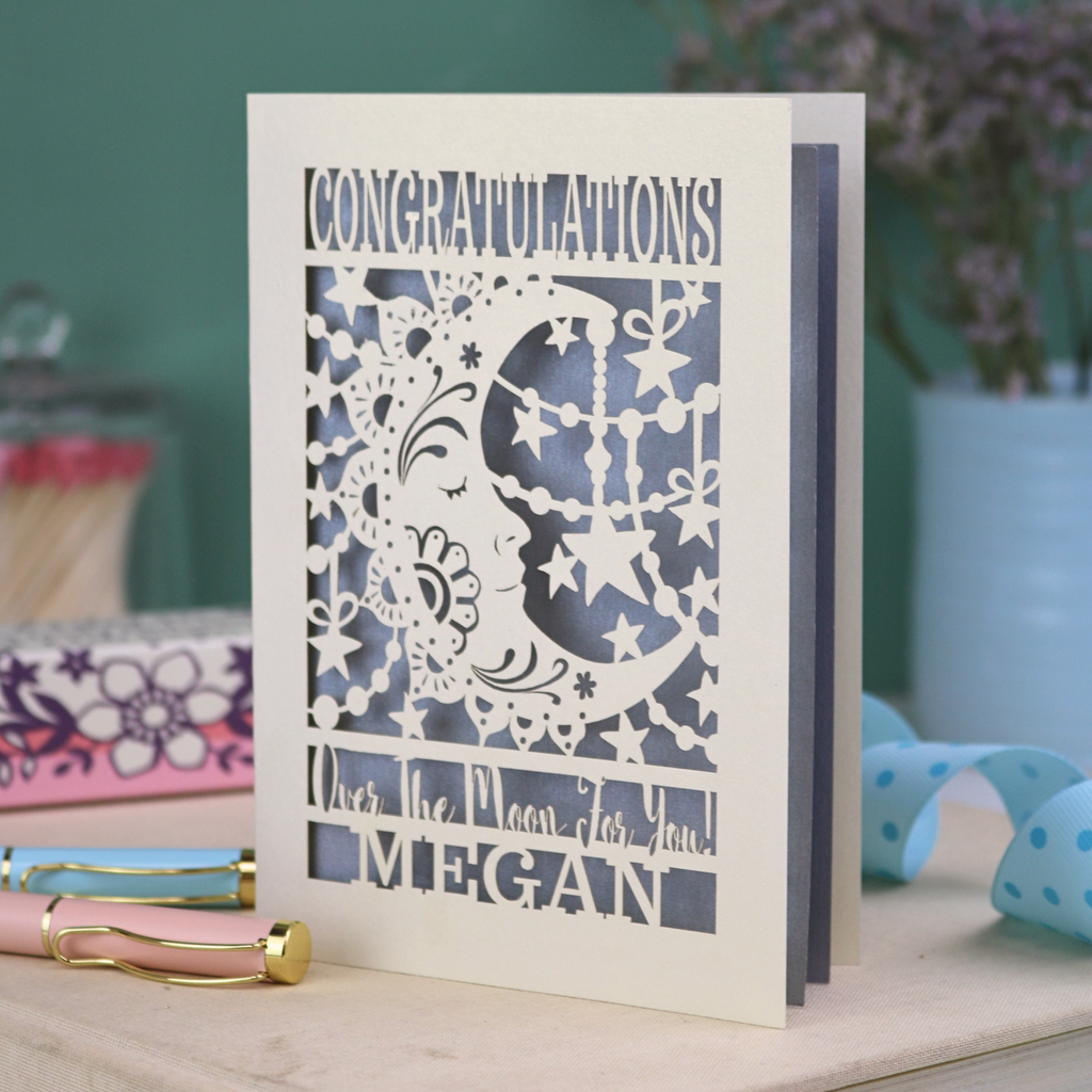 Personalised Congratulations Card with laser cut crescent moon design and text 'Congratulations, Over the moon for you Megan' on a wooden surface.