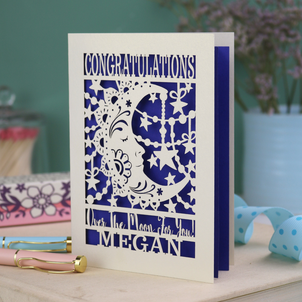 Personalised Congratulations Card with laser cut crescent moon design and text 'Congratulations, Over the moon for you Megan' on a wooden surface.