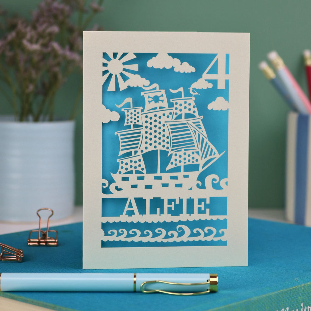 A personalised laser cut birthday card with cut out pirate ship illustration, the number 4 in the top right corner and the name Alfie 