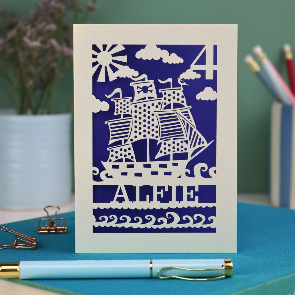 A personalised laser cut birthday card with cut out pirate ship illustration, the number 4 in the top right corner and the name Alfie 