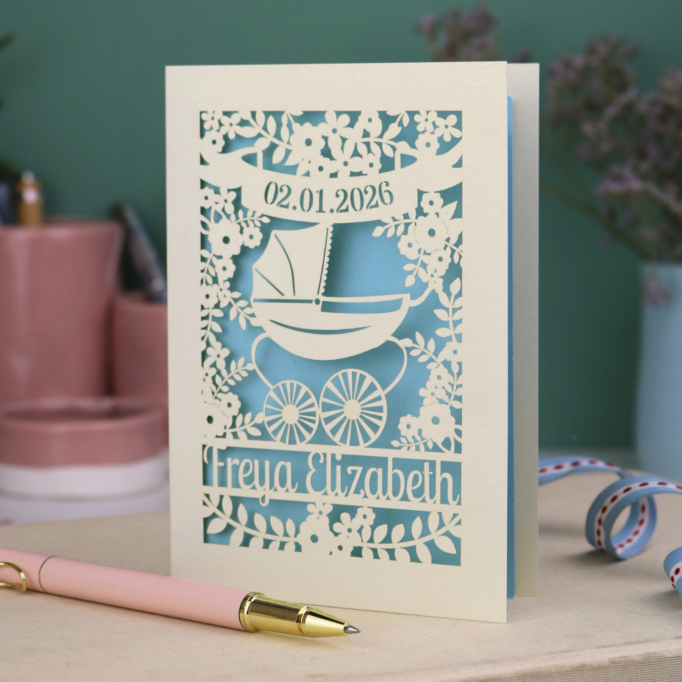 A personalised new baby card with laser cut floral vintage pram design. The baby's date of birth is cut out in stencil lettering in a banner, and the baby's name is at the bottom of the card.