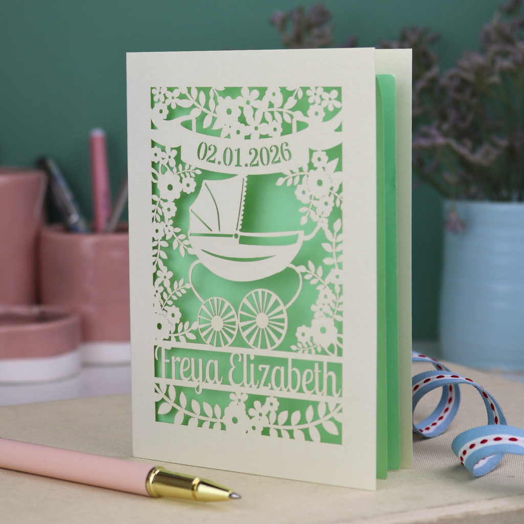 A personalised new baby card with laser cut floral vintage pram design. The baby's date of birth is cut out in stencil lettering in a banner, and the baby's name is at the bottom of the card.