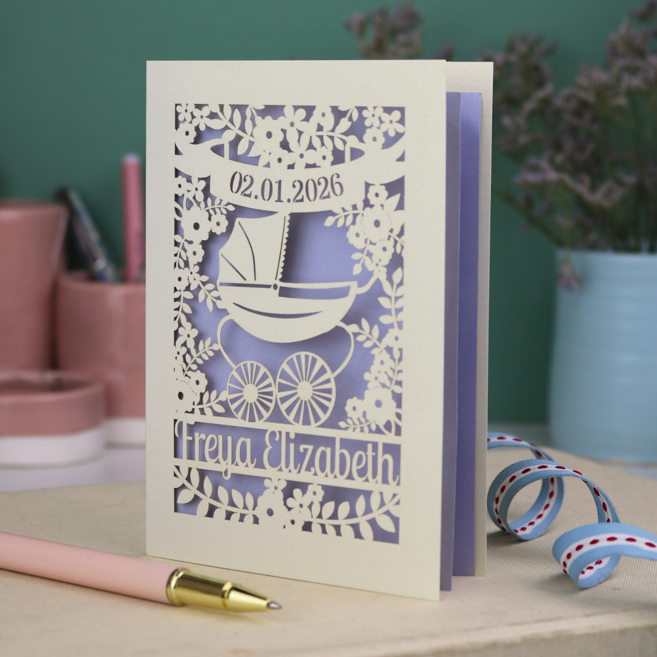 A personalised new baby card with laser cut floral vintage pram design. The baby's date of birth is cut out in stencil lettering in a banner, and the baby's name is at the bottom of the card.