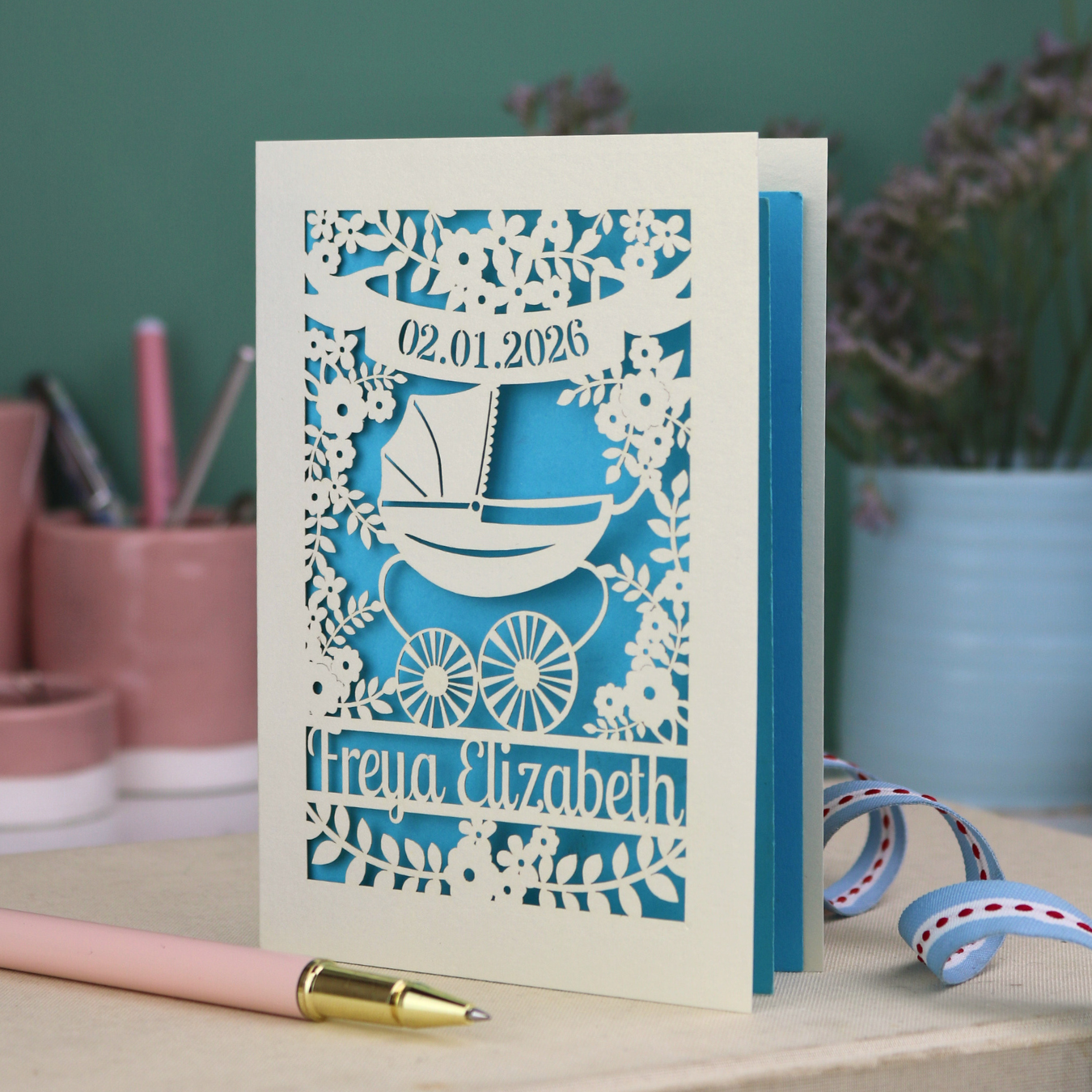 A personalised new baby card with laser cut floral vintage pram design. The baby's date of birth is cut out in stencil lettering in a banner, and the baby's name is at the bottom of the card.