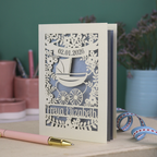A personalised new baby card with laser cut floral vintage pram design. The baby's date of birth is cut out in stencil lettering in a banner, and the baby's name is at the bottom of the card.
