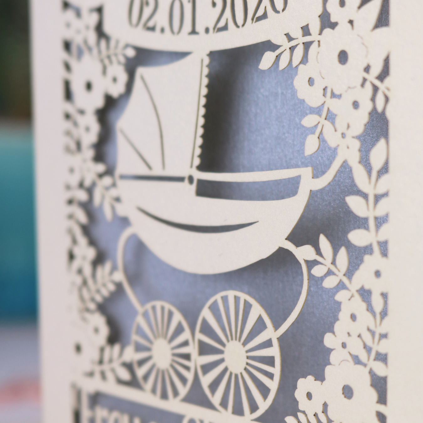 A personalised new baby card with laser cut floral vintage pram design. The baby's date of birth is cut out in stencil lettering in a banner, and the baby's name is at the bottom of the card.