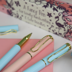 Two light blue ballpoint pens and a light pink ballpoint pen, all with gold accents on a pink surface, with a decorative box in the background.
