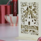 Decorative birthday card with castle design and 'Delilah' text on a table with red and white striped items.