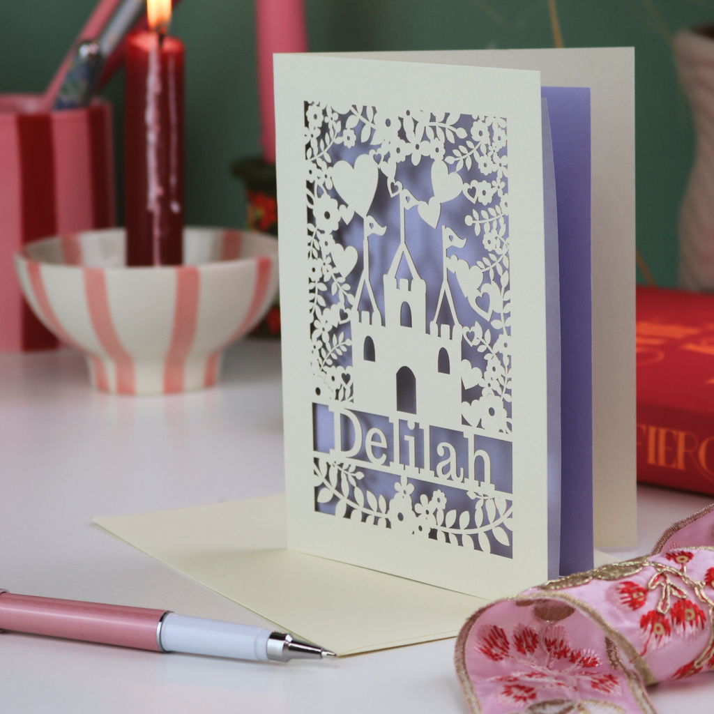 Laser-cut castle birthday card with 'Delilah' design on a table with stationery items.