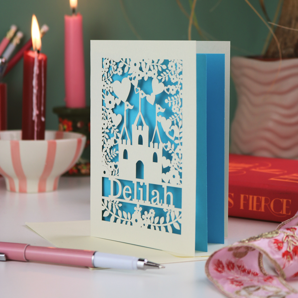 Decorative laser cut birthday card with cut-out design and 'Delilah' text, placed on a surface with candles and books in the background.