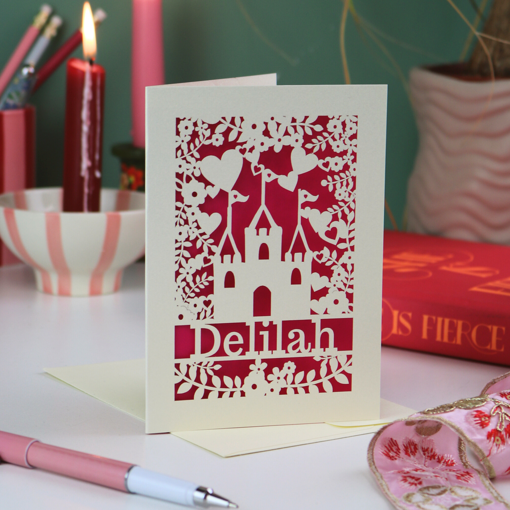 Decorative greetings card with castle design and 'Delilah' text on a table with candles and books.