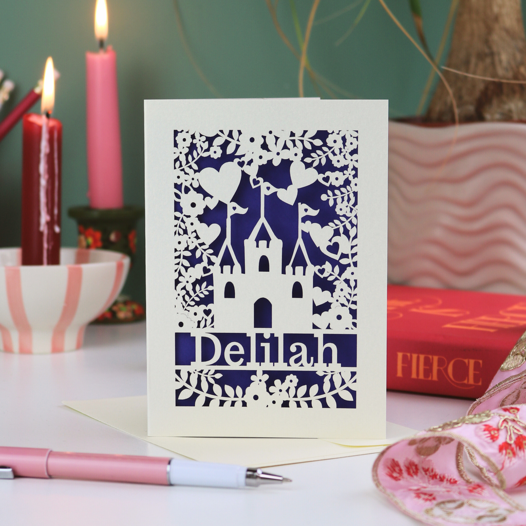 Decorative birthday card with cut-out design and 'Delilah' text, placed on a surface with candles and books in the background.