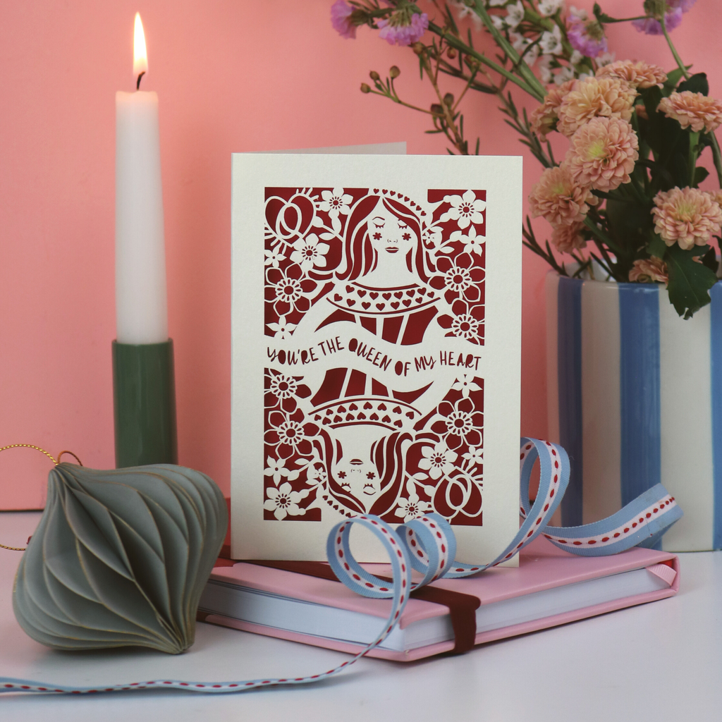 Decorative Queen of Hearts laser cut card with floral design and text, placed on books with a candle and flowers in the background.