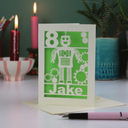 A laser cut robot birthday card with cut out design, age 8 in the top left corner and 'Jake' at the bottom. Card is surrounded by candles, plants and a pen