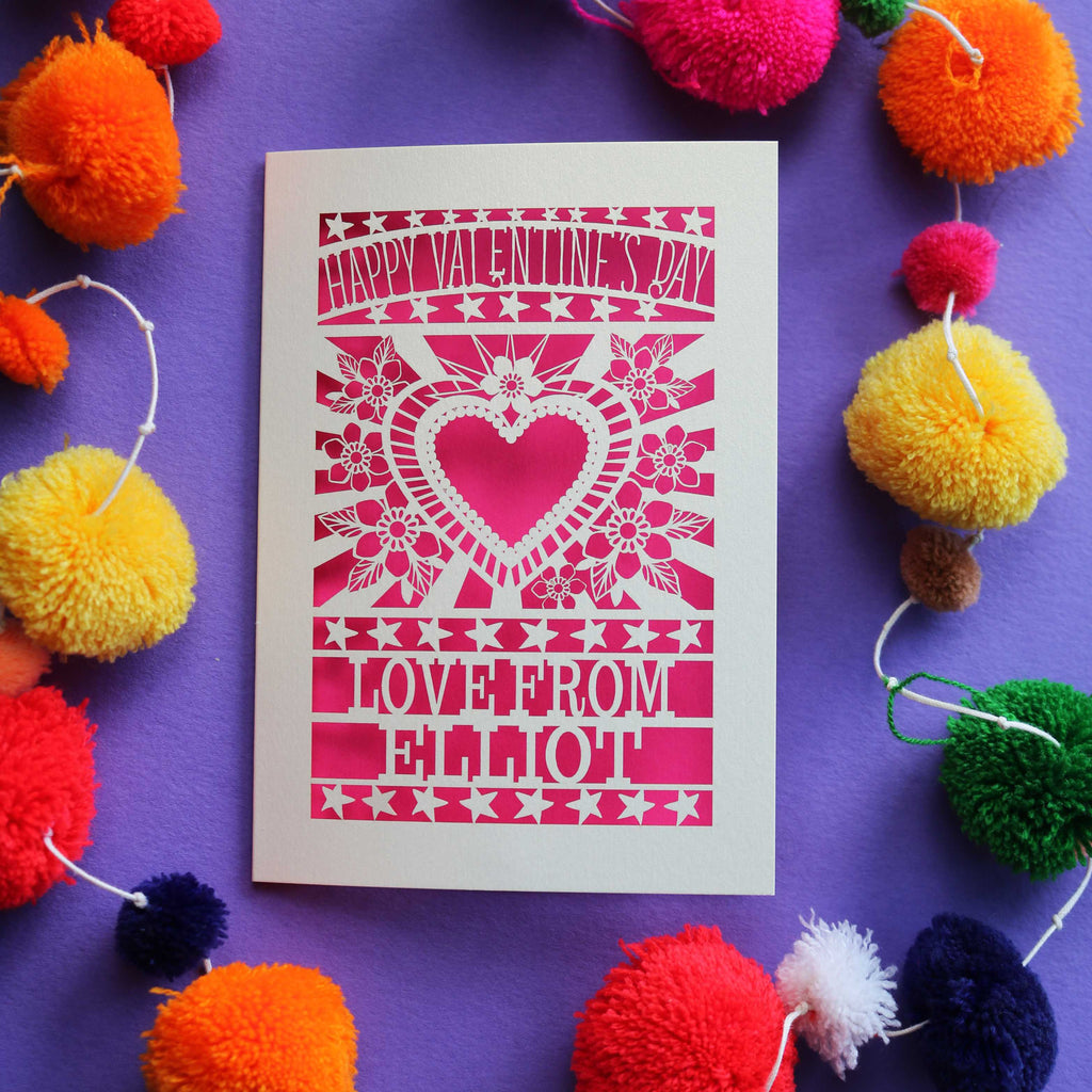 A laser cut personalised Valentines card that has a heart and flowers cut out and text that says "Happy Valentine's Day, love from" and a name
