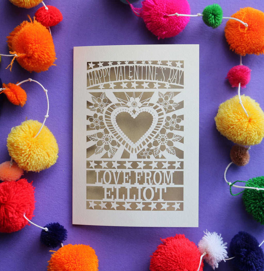 A laser cut personalised Valentines card that has a heart and flowers cut out and text that says "Happy Valentine's Day, love from" and a name