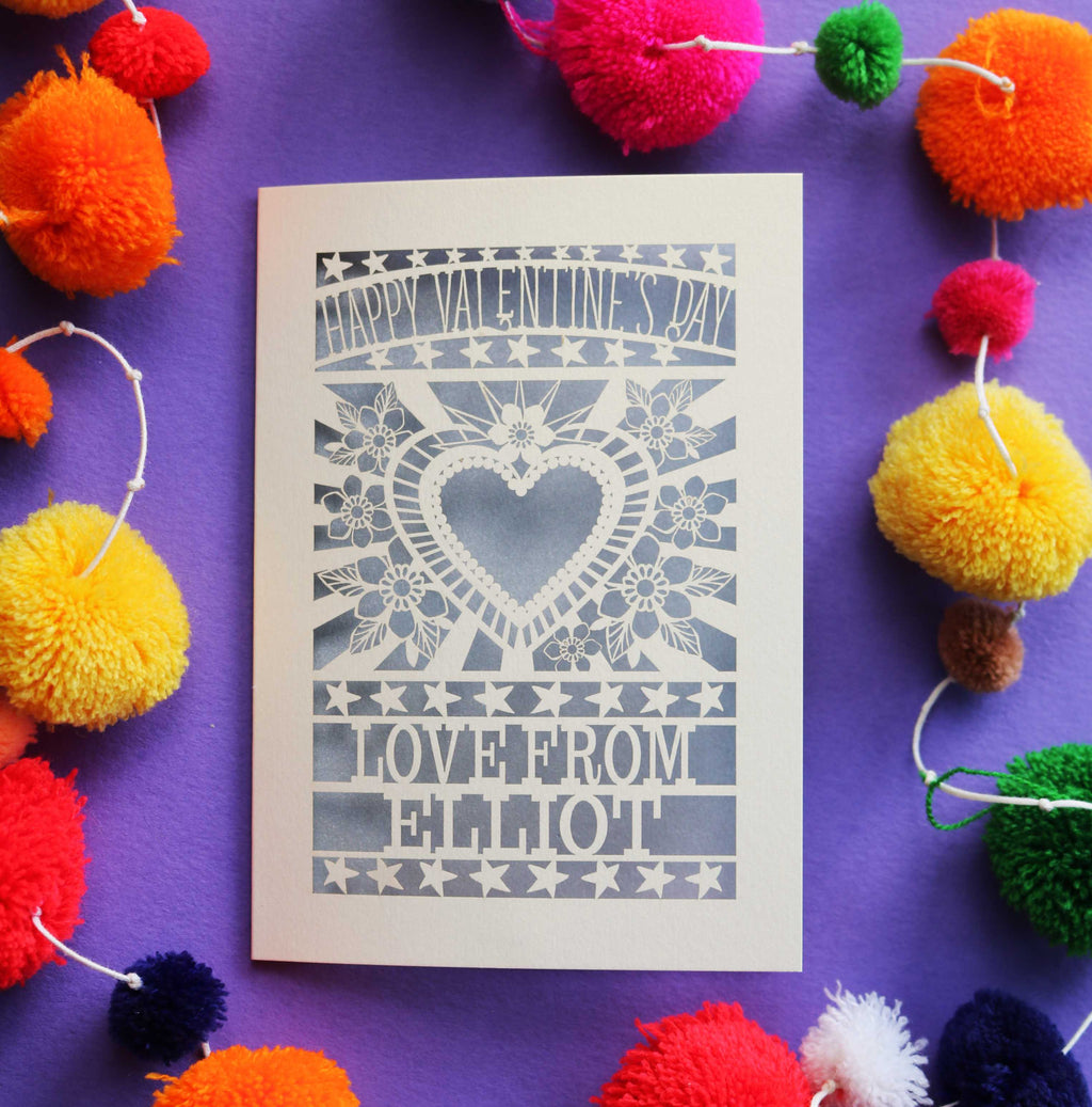 A laser cut personalised Valentines card that has a heart and flowers cut out and text that says "Happy Valentine's Day, love from" and a name