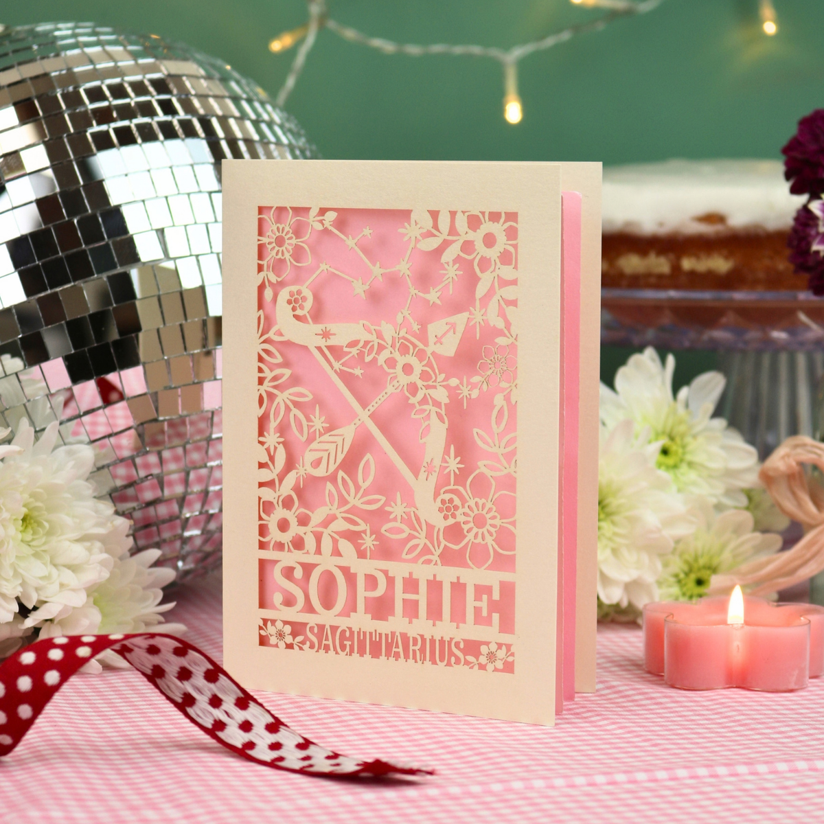 A personalized laser-cut birthday card with a pink and cream design, featuring the name 'SOPHIE' and the zodiac 'SAGITTARIUS', styled on a pink checked tablecloth with a lit candle, white flowers, a disco ball, and string lights in the background.