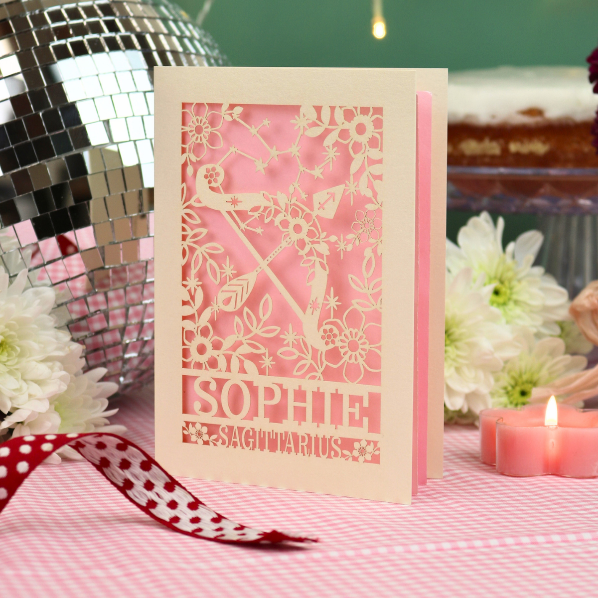 A personalized laser-cut greeting card for the Sagittarius zodiac sign 'Sophie', featuring a pink and cream design with a celestial motif, displayed on a pink checkered tablecloth with a red polka dot ribbon, white flowers, and a disco ball in the background.