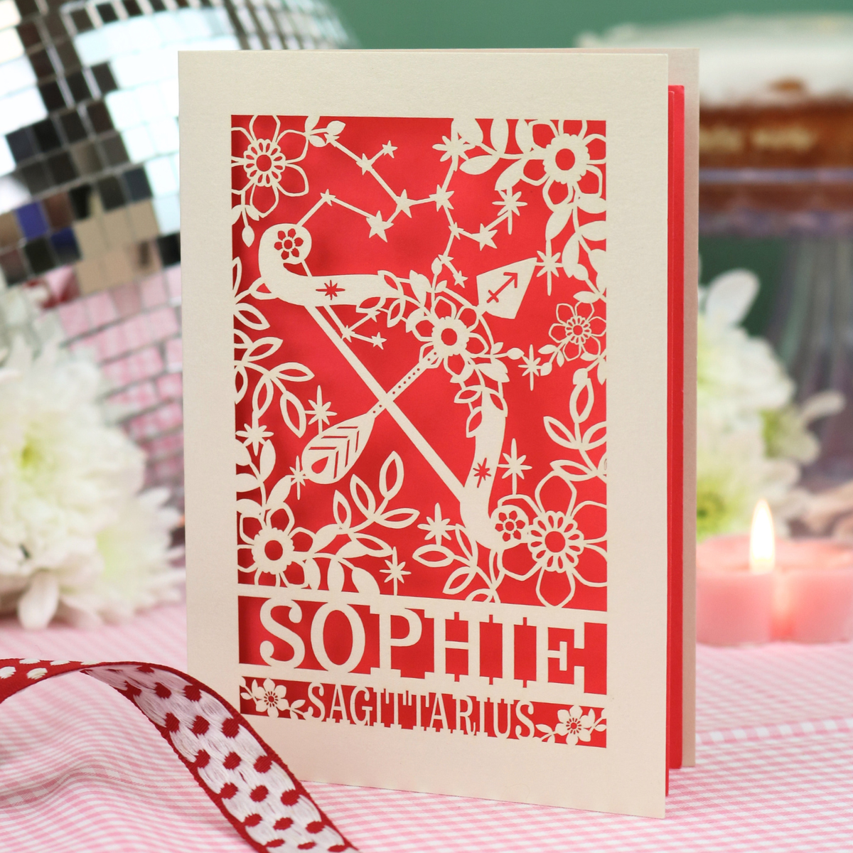 A laser-cut Sagittarian birthday card featuring a bow and arrow, floral patterns, and the name 'SOPHIE' with 'SAGITTARIUS' at the bottom. The card is displayed on a checked tablecloth with a red and white polka dot ribbon in the foreground and a blurred background with flowers and a candle.