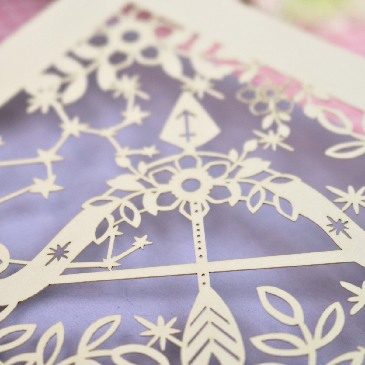 Close-up of a laser-cut decorative paper artwork featuring a bow and arrow motif with floral and constellation details, layered over a lavender-purple background.