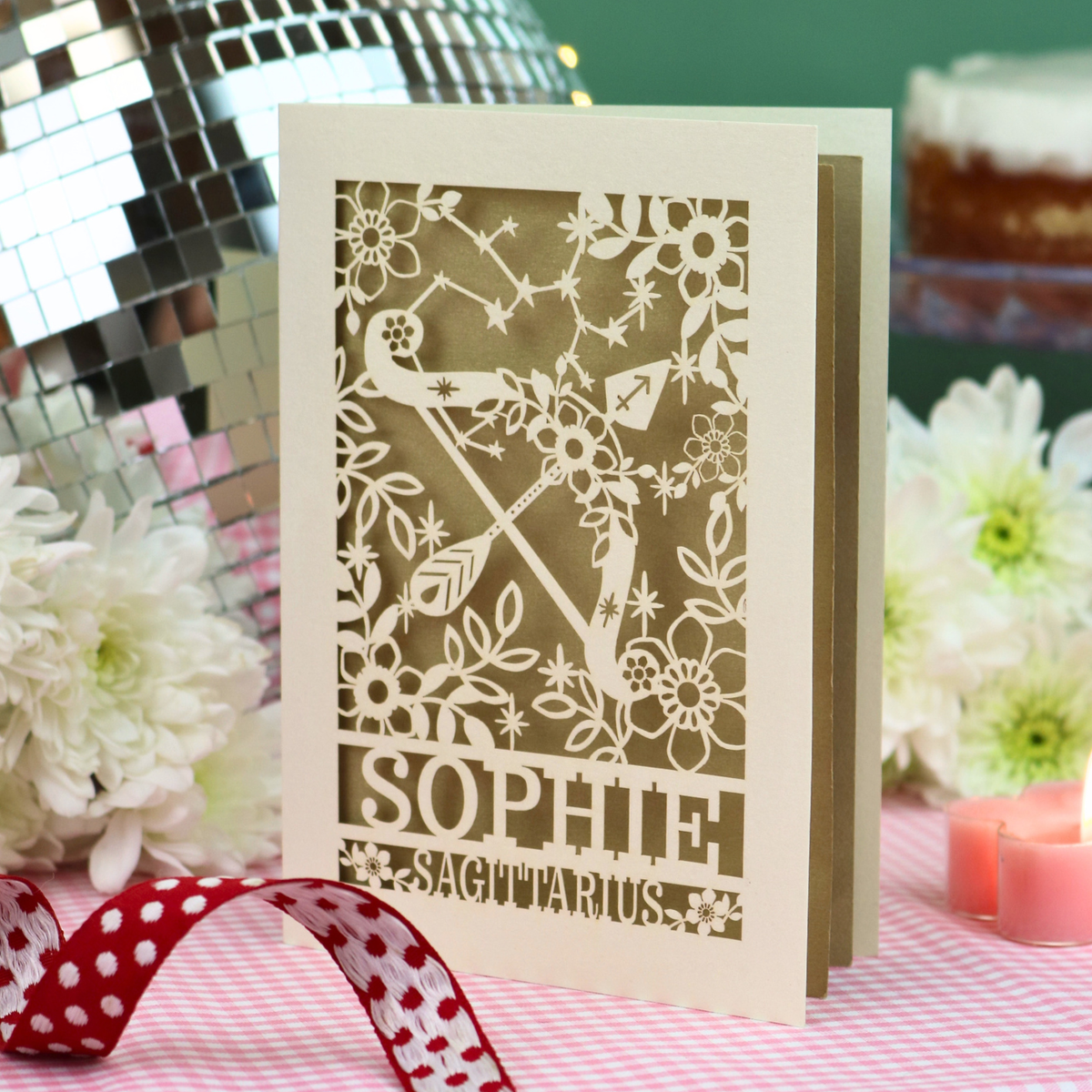 A handmade birthday card with a laser-cut design featuring a bird, floral patterns, and the text 'SOPHIE SAGITTARIUS', displayed on a pink-and-white checked tablecloth with a red polka-dot ribbon, white flowers, candles, and a disco ball in the background.
